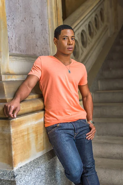 Dressing in a light orange short sleeve V neck shirt, jeans,  a young handsome black student is standing downstairs against a column, charmingly looking at you