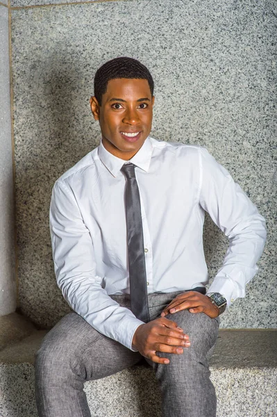 Beyaz gömlekli, siyah pantolonlu, elleri kucağında dinlenen, taş bir bankta oturan yakışıklı siyah bir üniversite öğrencisi, sana gülümseyerek bakıyor.