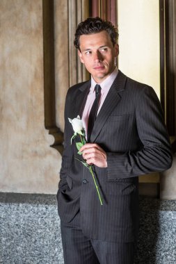 Pencerenin yanında duran ve resmi olarak giyinen genç yakışıklı bir adam elinde beyaz bir gül tutuyor ve seni bekliyor.