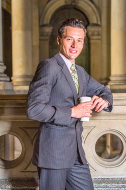 Resmi giyinmiş, elinde bir fincan kahve olan genç bir iş adamı, ofis koridorunda korkulukların önünde durmuş, sana gülümsüyor ve gülümsüyor.