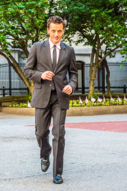 Siyah çizgili ceket, pantolon, kravat, deri ayakkabı giyen genç bir işadamı ofis binasının önünde yürüyor.