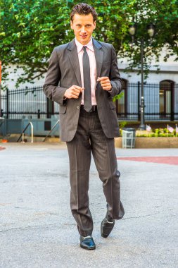 Siyah çizgili ceket, pantolon, kravat, deri ayakkabı giyen genç bir işadamı ofis binasının önünde yürüyor.