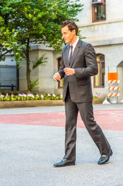 Siyah çizgili ceket, pantolon, kravat, deri ayakkabı giyen genç bir işadamı ofis binasının önünde yürüyor.