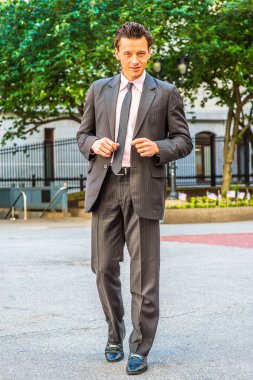 Siyah çizgili ceket, pantolon, kravat, deri ayakkabı giyen genç bir işadamı ofis binasının önünde yürüyor.