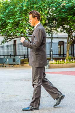 Siyah çizgili ceket, pantolon, kravat, deri ayakkabı giyen genç bir işadamı ofis binasının önünde yürüyor.