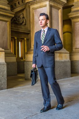 Resmi olarak takım elbise, kravat ve deri ayakkabı giyen, deri çanta taşıyan ve cep telefonu tutan genç bir işadamı, bir iş binasının önünden geçiyor.