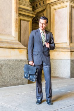 Resmi olarak takım elbise, kravat ve deri ayakkabı giyen, deri çanta taşıyan genç bir işadamı eski moda bir iş binasında sizi bekliyor.