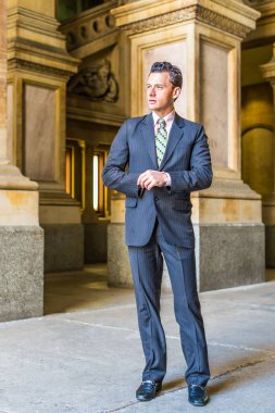 Resmi olarak takım elbise, kravat ve deri ayakkabılar giyen genç bir işadamı, eski moda bir iş binasının içinde duruyor ve kendinden emin bir şekilde ileriye bakıyor.