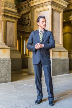 Resmi olarak takım elbise, kravat ve deri ayakkabılar giyen genç bir işadamı, eski moda bir iş binasının içinde duruyor ve kendinden emin bir şekilde ileriye bakıyor.