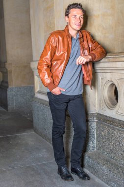 Kahverengi deri ceket, gri atlet, siyah pantolon, bir elini cebine sokmuş genç yakışıklı bir adam koridorda korkuluk gibi duruyor, gülümsüyor ve düşünüyor.