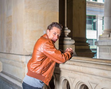 Kahverengi deri ceket, gri atlet, siyah pantolon, genç yakışıklı bir adam balkonda çitin yanında duruyor, başı dönüyor, gülümsüyor ve sana bakıyor.