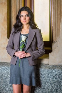 Gri örgü bir elbise ve ceket giymiş, elinde beyaz bir çiçek tutan genç bir iş kadını, küçük bir pencerenin kenarında durmuş, samimi bir şekilde savaş arıyor.