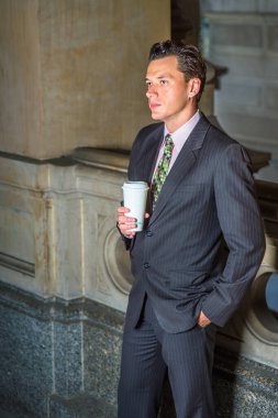 Resmi olarak giyinen, bir elini cebine koyan ve bir elini bir fincan kahve tutan genç bir işadamı, ofis koridorunda dikilip, derin düşüncelere dalmış,.