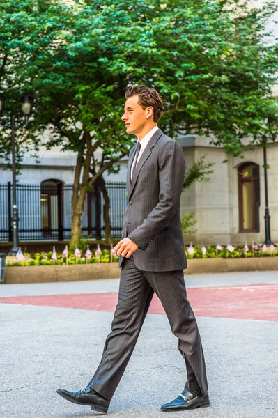 Siyah çizgili ceket, pantolon, kravat, deri ayakkabı giyen genç bir işadamı ofis binasının önünde yürüyor.