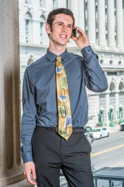 Mavi gömlekli, siyah pantolonlu, renkli kravatlı genç bir üniversite öğrencisi bir iş binasının önünde sırıtarak bekliyor.