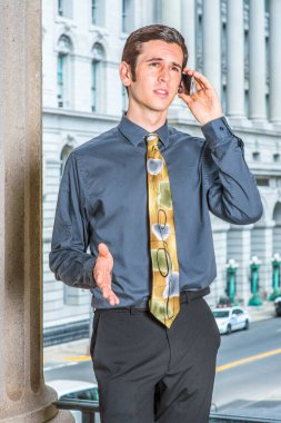 Mavi gömlekli, siyah pantolonlu, renkli kravatlı genç bir işadamı bir iş binasının önünde duruyor, bir elini hareket ettiriyor ve kaşlarını çatıyor, bir telefon görüşmesi yapıyor.