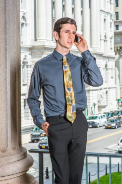 Mavi gömlekli, siyah pantolonlu, renkli kravatlı genç bir işadamı bir iş binasının önünde duruyor, kaşlarını çatmış, telefonda konuşuyor.