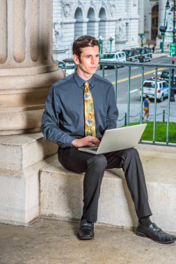 Mavi gömlekli, siyah pantolonlu, renkli kravatlı genç bir iş adamı bir iş binasının önünde oturmuş kucağında bilgisayarla çalışıyor.