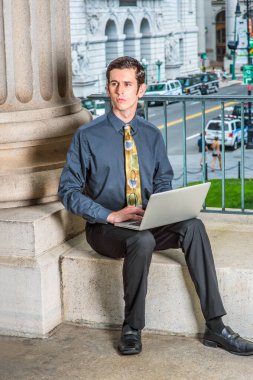 Mavi gömlekli, siyah pantolonlu, renkli kravatlı genç bir üniversite öğrencisi kampüsün dışında oturmuş kucağında bilgisayarla çalışıyor.