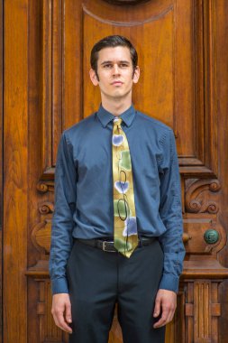 Mavi gömlekli, pantolonlu ve renkli kravatlı genç bir üniversite öğrencisi eski moda ofis kapısının önünde dikiliyor.