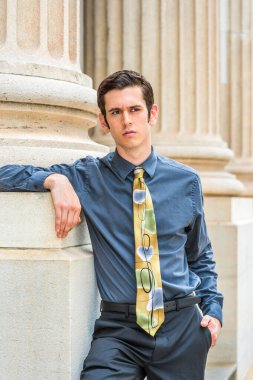 Mavi gömlekli, siyah pantolonlu, renkli kravatlı genç bir üniversite öğrencisi bir ofis binasının önünde dikiliyor.