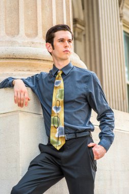 Mavi gömlekli, siyah pantolonlu, renkli kravatlı genç bir üniversite öğrencisi ofis binasının önünde bekliyor.