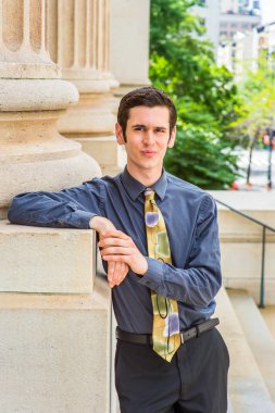 Mavi gömlekli, siyah pantolonlu, renkli kravatlı genç bir üniversite öğrencisi bir ofis binasının önünde durmuş sana bakıyor.