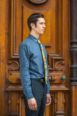 Mavi gömlekli, pantolonlu ve renkli kravatlı genç bir üniversite öğrencisi eski moda ofis kapısının önünde dikiliyor.