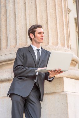Resmi olarak siyah takım elbise giymiş, siyah kravat takmış genç bir iş adamı ofis binasının önünde bilgisayar üzerinde çalışıyor.