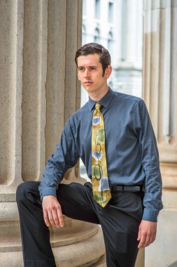 Mavi gömlekli, siyah pantolonlu, renkli kravatlı, bir kolu bacağının üzerinde dinlenen genç bir üniversite öğrencisi, bir ofis binasının dışında, derin düşüncelere dalmış bir şekilde duruyor.