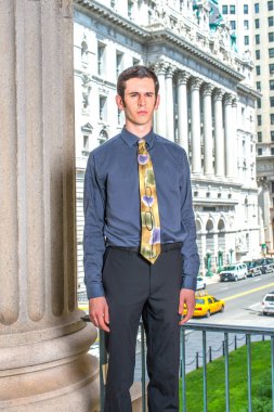 Mavi gömlekli, siyah pantolonlu, renkli kravatlı genç bir üniversite öğrencisi bir ofis binasının önünde duruyor.