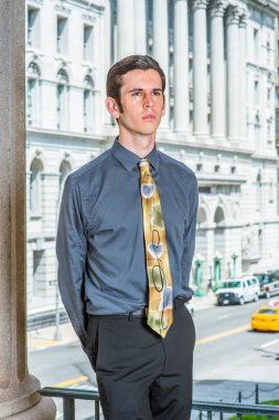 Mavi gömlekli, siyah pantolonlu, renkli kravatlı, ellerini arkaya koyan genç bir üniversite öğrencisi bir ofis binasının önünde dinleniyor ve düşünüyor.