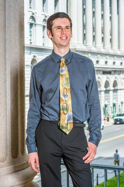 Mavi gömlekli, siyah pantolonlu, renkli kravatlı genç bir üniversite öğrencisi bir iş binasının önünde sırıtarak bekliyor.