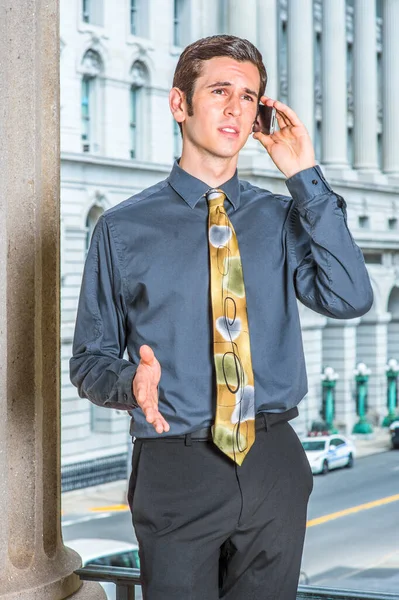 Mavi gömlekli, siyah pantolonlu, renkli kravatlı genç bir işadamı bir iş binasının önünde duruyor, bir elini hareket ettiriyor ve kaşlarını çatıyor, bir telefon görüşmesi yapıyor.