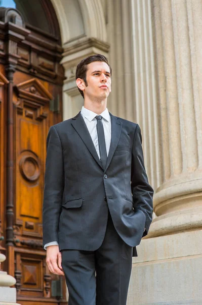 Resmi olarak siyah bir takım elbise, siyah bir kravat, bir elini cebine sokan genç bir işadamı bir ofis binasının önünde kendinden emin bir şekilde bekliyor.