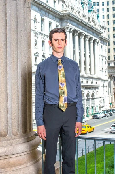 Mavi gömlekli, siyah pantolonlu, renkli kravatlı genç bir üniversite öğrencisi bir ofis binasının önünde duruyor.