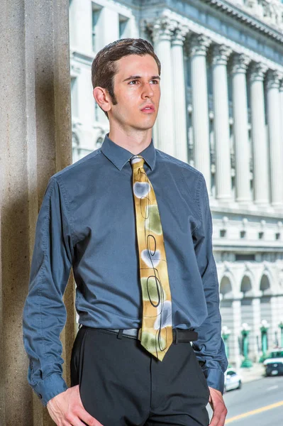 Mavi gömlekli, siyah pantolonlu, renkli kravatlı genç bir işadamı bir ofis binasının önünde kendinden emin bir şekilde bekliyor.