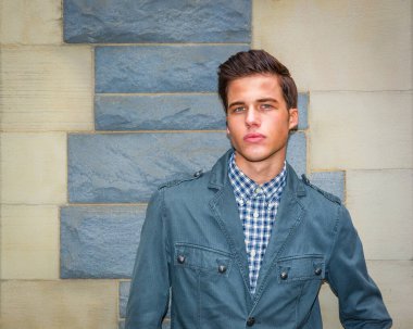 Şık Mavi Ceketli Adam. Saçları geriye taranmış bir adam taş bir duvara karşı poz veriyor. Damalı bir gömleğin üzerine mavi bir ceket giyer, modern ve modaya uygun bir görünüm verir.