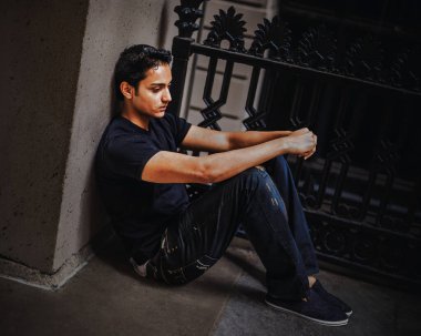 Yalnız oturan düşünceli genç adam. Sıradan bir kot pantolon ve tişört giymiş bir adam yere oturup duvara yaslanıyor. Loş ışıkla çevrili, dalgın dalgın ileri bakıyor.. 