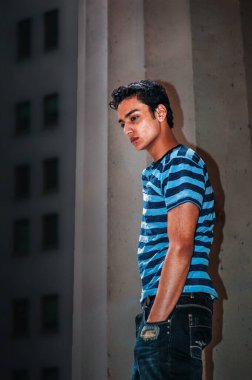 Çizgili Gömlekli Düşünceli Genç Adam. Mavi çizgili gömlekli genç bir adam elleri ceplerinde bir duvara yaslanıyor. Arkaplan kompozisyona derinlik ekleyen dikey çizgiler içeriyor.