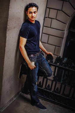 Duvara yaslanan genç adam. Genç bir adam bir ayağı duvara dayalıyken gelişigüzel ayakta duruyor. Koyu renk tişört ve kot pantolon giymiş, şehir ortamında..