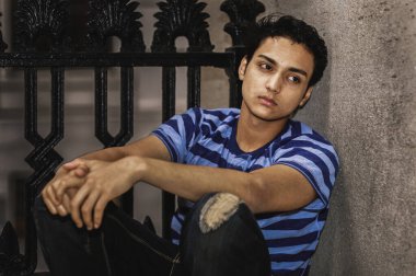 Çizgili gömlekli dalgın genç adam. Genç bir adam, mavi ve siyah çizgili bir gömlek giymiş, dokulu taştan bir duvarın karşısında oturuyor. Uzağa dikkatlice bakar, ellerini sıkarak..