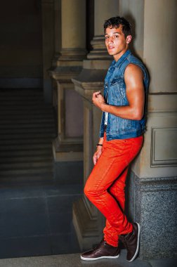 Şehir Ayarları 'nda gündelik moda. Genç bir adam kendinden emin bir şekilde kaya çıkıntısına karşı duruyor. Kot yelek ve parlak kırmızı pantolon giyiyor, sessiz mimari arka plana karşı koyu bir kontrast oluşturuyor.