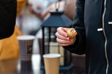 Elinde bisküvi tutan bir adam, arka planda kağıt bardaklı açık bir kafeterya masası ve bir fener, yakın plan.