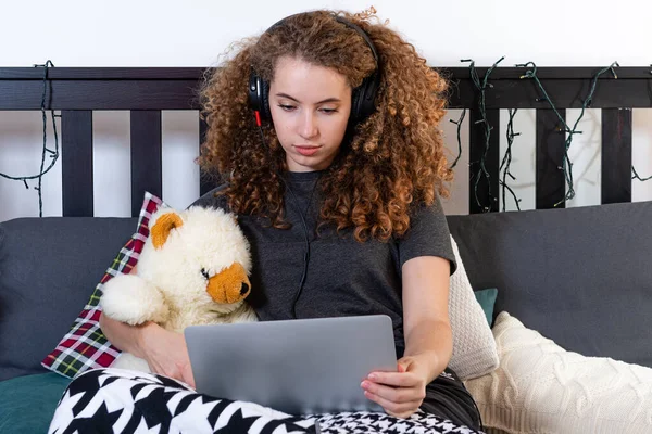 a young curly teen with a teddy bear sits on the bed and watches a movie on her laptop