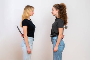 İki kızgın kadın karşı karşıya duruyor ve sırtlarında bıçak ve balta saklıyorlar, beyaz arka planda izole edilmiş, gizli nefret ve ikiyüzlülük kavramı.