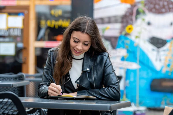 屋外のカフェに座っている間にノートにメモを取る長い髪を持つ素晴らしい楽観的な若い女性 — ストック写真