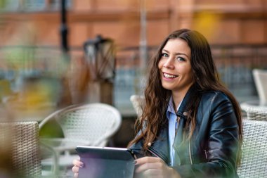 Sokak kafeteryasında oturmuş kameraya bakarken tabletle çalışan genç bir kadın.