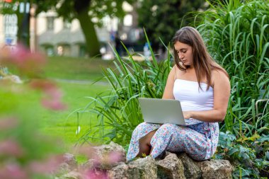 Parkta oturan genç bir kadın dizüstü bilgisayar, uzaktan öğrenme ve çalışma konsepti üzerinde çalışıyor.
