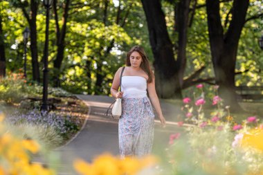 Açık bluz giyen ve değişik etek giyen genç bir kadın şehir parkında yürüyor, ön plandaki çiçekleri dağıtıyor.